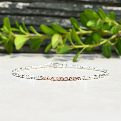 A delicate beaded bracelet with alternating silver and rose gold beads, displayed on a light beige surface with blurred green leaves in the background.