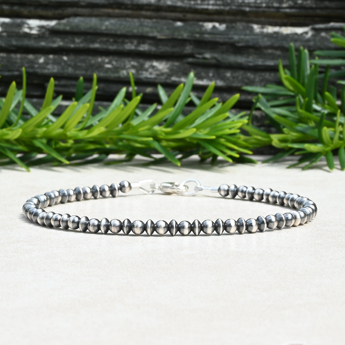 A sterling silver beaded bracelet with faceted saucer-shaped beads, displayed on a light beige surface with green pine branches and a dark wood background.