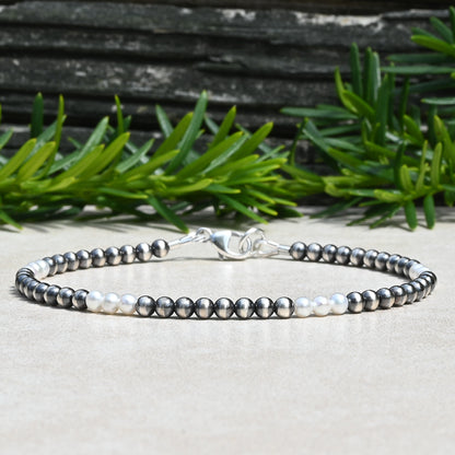 A beaded bracelet with alternating dark gray and white pearls, featuring a silver lobster clasp, displayed on a light beige surface with green foliage and a dark wood background.