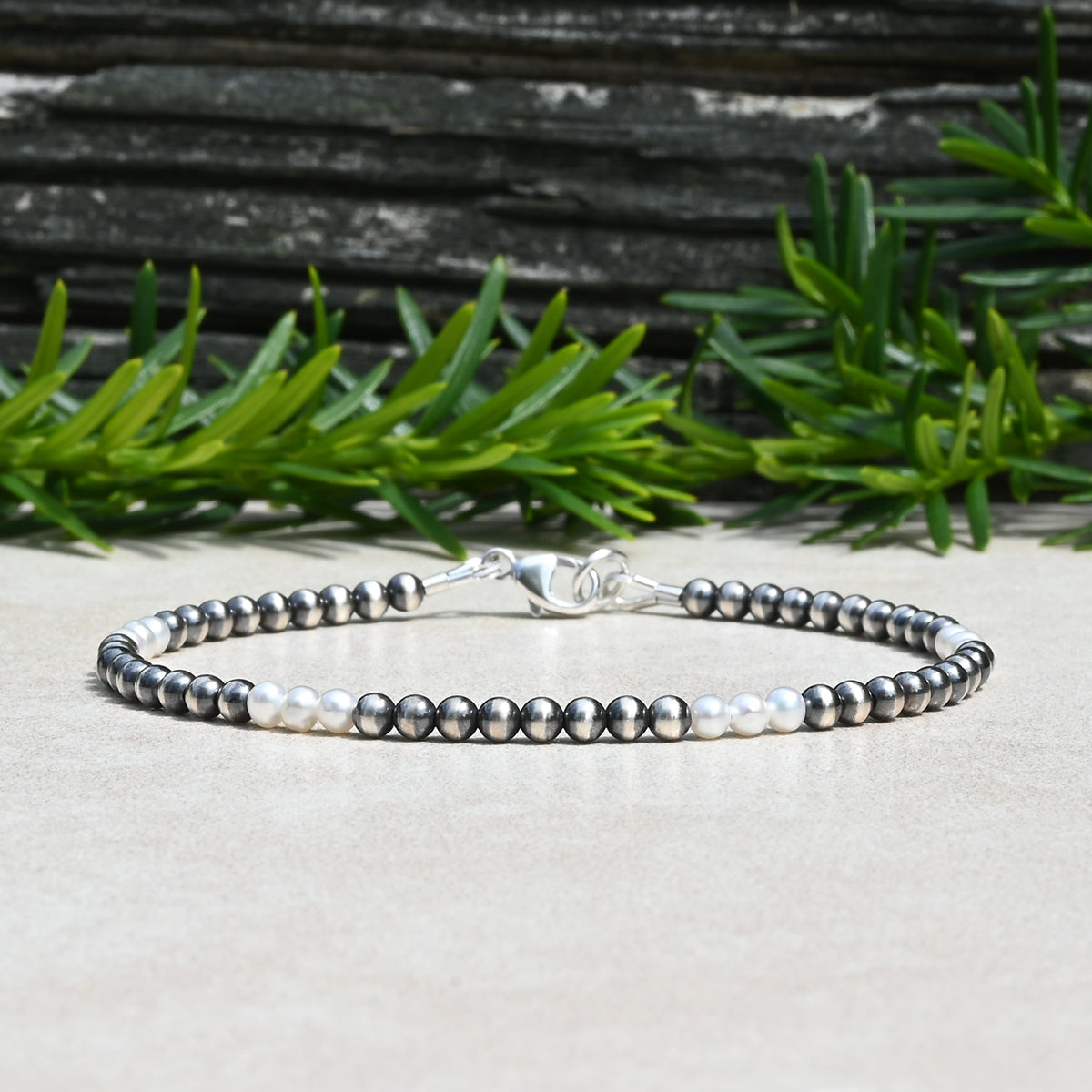 A Sterling Silver and Freshwater Pearl beaded bracelet with alternating dark gray and white pearls, secured with a silver clasp, displayed on a light beige surface with green foliage and a dark textured background.