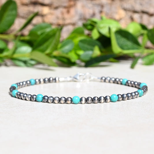 A beaded bracelet with alternating metallic silver-gray and bright turquoise blue beads, displayed on a light surface with a blurred background of green leaves and wood.